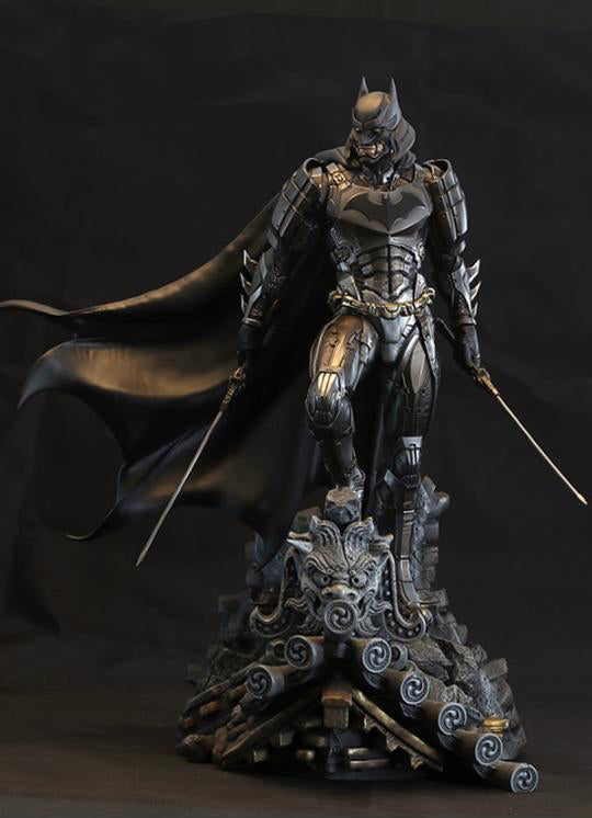 A detailed Batman armored statue in dark metallic silver and black, standing on a decorative base with swords in both hands against a dark studio background.