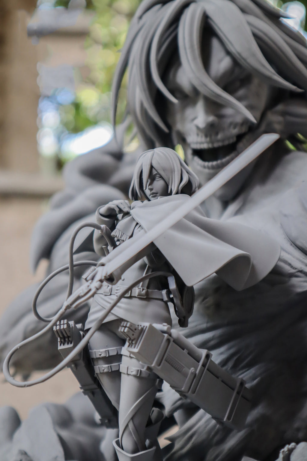 A close-up of a gray collectible figurine depicting a female tactical character holding a sword in front of a large skull-faced monster, with a blurred outdoor background.