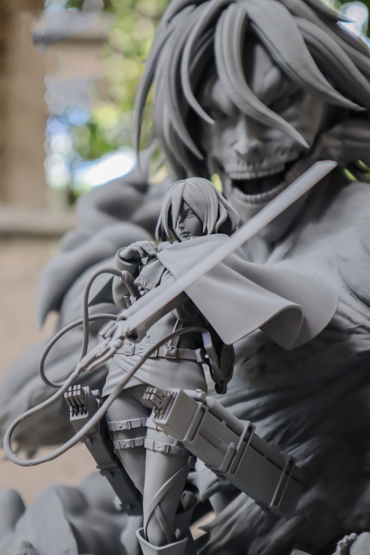 A close-up of a gray collectible figurine depicting a female tactical character holding a sword in front of a large skull-faced monster, with a blurred outdoor background.