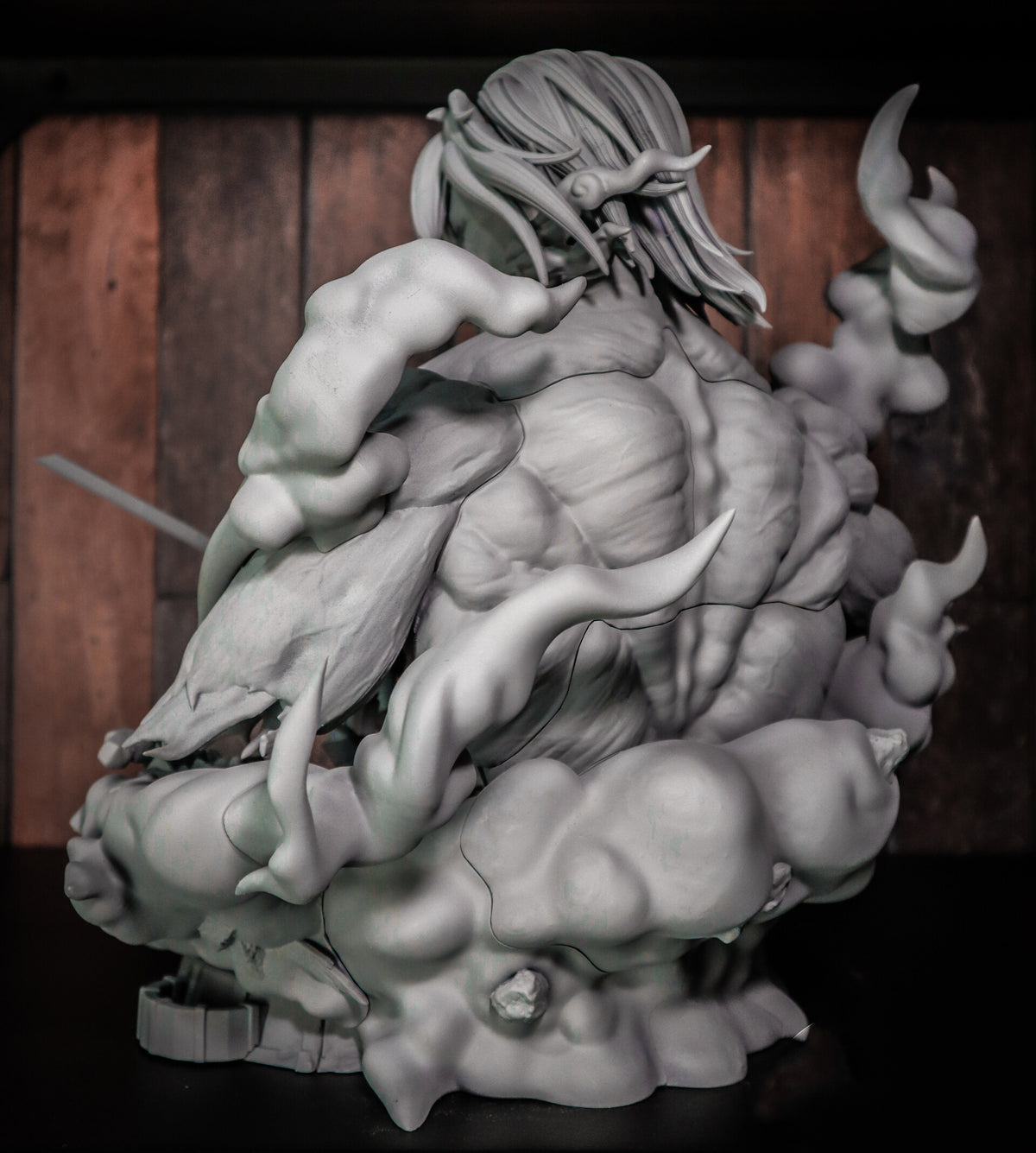 A detailed 3D-printed or sculpted fantasy character figurine shown in unpainted light gray resin/plastic. The character has long flowing hair, muscular torso details, and is posed amidst swirling smoke or cloud-like forms, set against a dark wooden background.