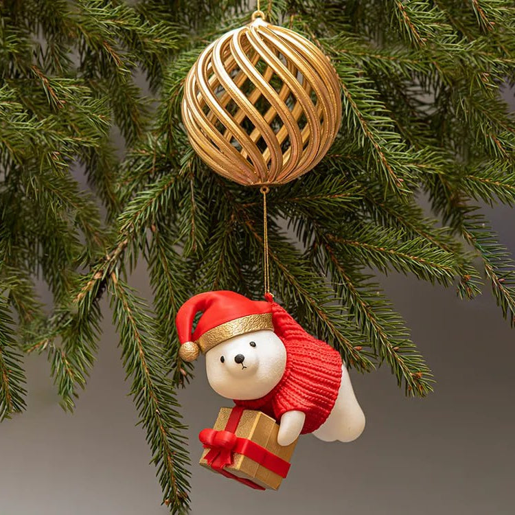 Christmas ornament of a polar bear in a red sweater with a gift, hanging from a gold ball on a tree branch.
