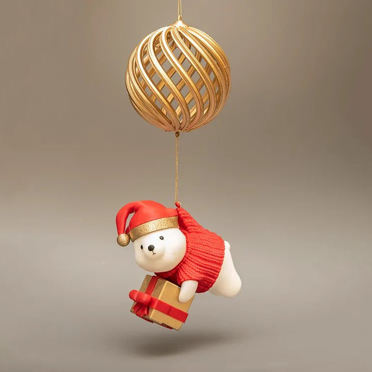 Decorative ornament with a white bear in a red outfit and Santa hat, holding a gift, hanging from a gold ball on a beige background.
