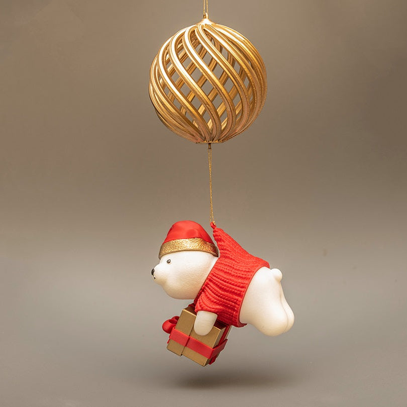 Decorative ornament of a bear in a red outfit with a gold gift box, hanging from a gold ball on a beige background.