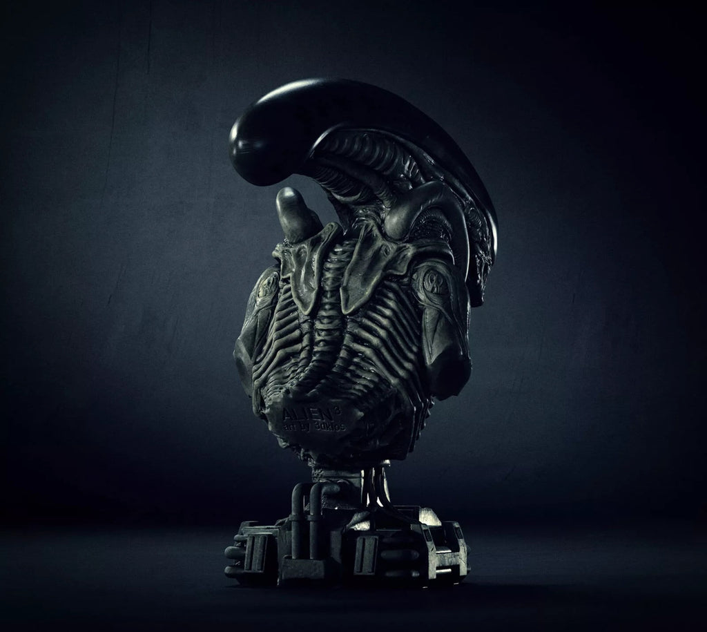 A dark metallic sci-fi bust sculpture of an Alien Xenomorph torso on a pedestal, photographed against a moody dark gray studio background.