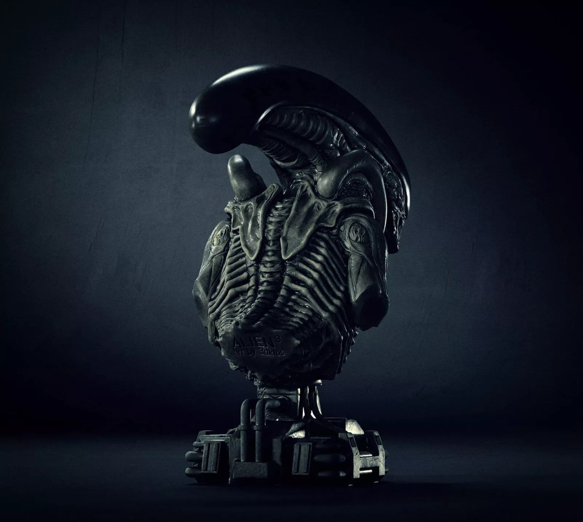 A dark metallic sci-fi bust sculpture of an Alien Xenomorph torso on a pedestal, photographed against a moody dark gray studio background.