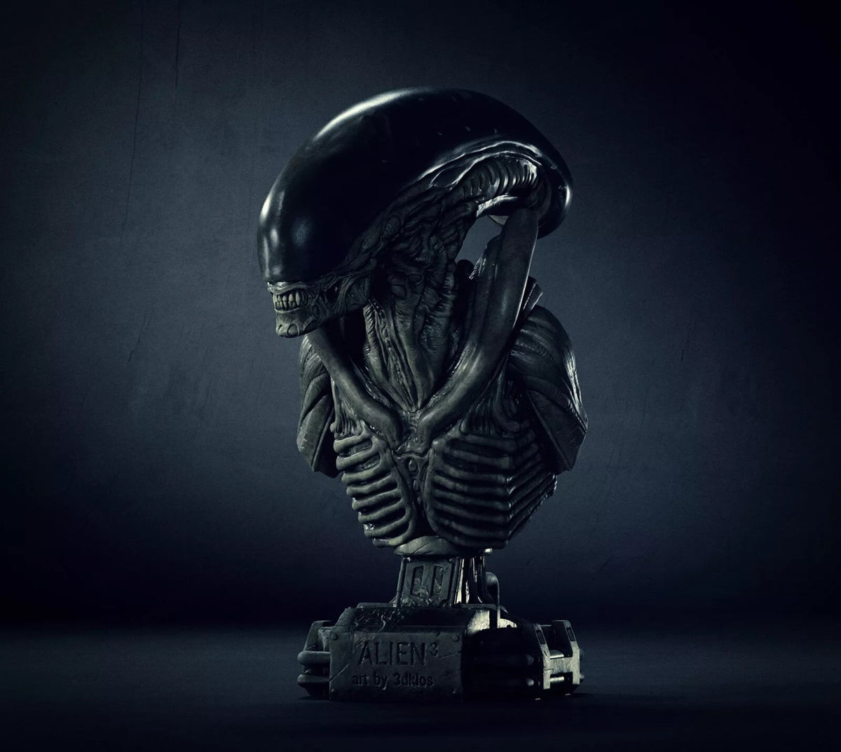 A dark metallic bust sculpture of an Alien Xenomorph on a pedestal base, photographed against a moody dark gray studio background.