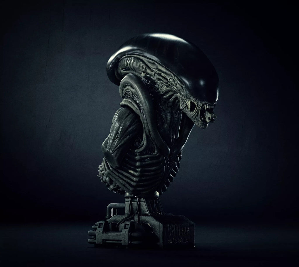 A dark, highly detailed bust sculpture of an Alien Xenomorph on a mechanical base, photographed against a very dark studio background.