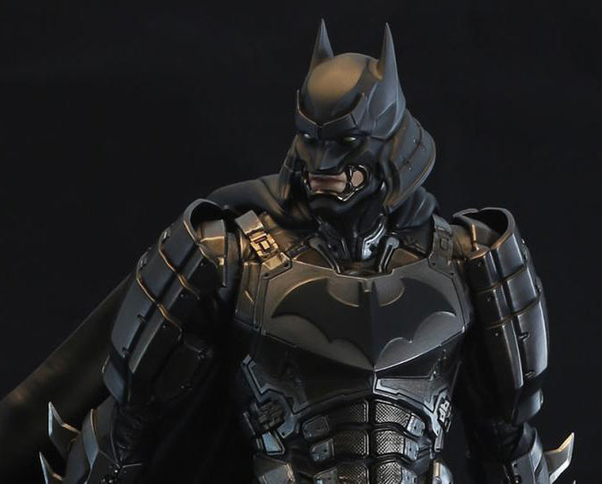 A close-up studio shot of a Batman armored figure or collectible bust against a dark black background. The character wears detailed dark gray and black futuristic bat armor with a cowl and cape visible at the sides.
