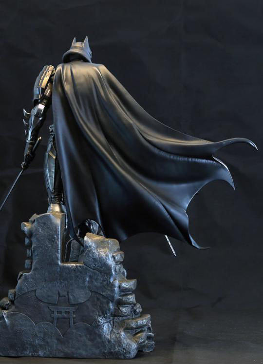A Batman statue shown from the back, wearing a flowing black cape and armored suit, standing on a dark rocky base against a black backdrop.