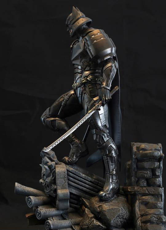 A dark metallic Batman armored figure posed on a rocky base with a sword, photographed against a black studio background.