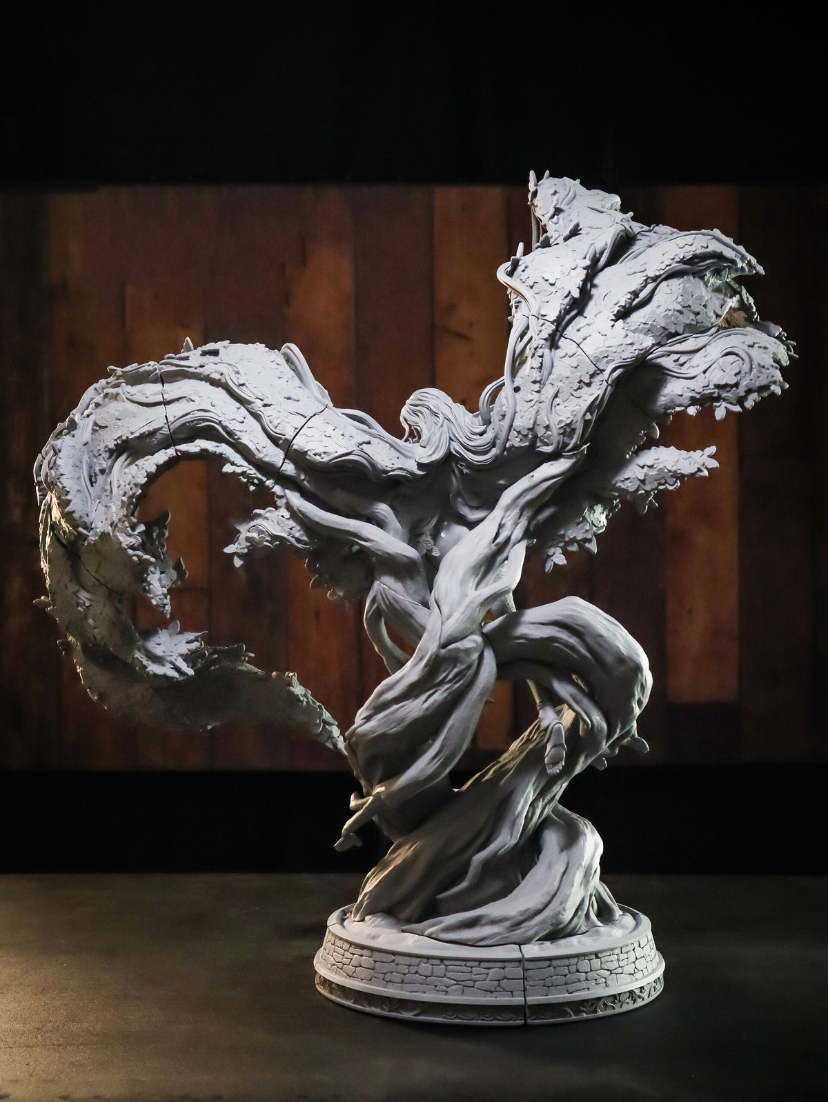 A detailed fantasy sculpture of a swirling, wind-blown tree trunk or elemental form on a circular base, photographed against a dark wood-paneled background.