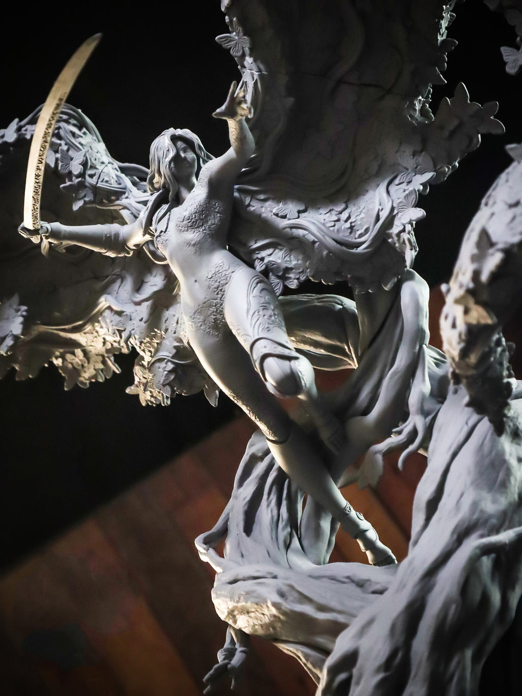A detailed fantasy sculpture of a winged female figure with a curved sword, shown in a monochrome gray material against a dark background.